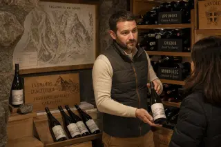Caviste presenting an unbranded wine bottle in a tidy alpine cellar with warm, subdued lighting and neatly arranged shelves.