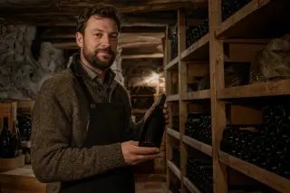 caviste in a warmly lit alpine wine cellar presenting an unlabeled bottle, surrounded by neatly arranged racks and stone walls
